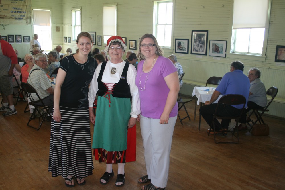 Marianne Marttila-Klipfel (whose photography is on display in the background), Annikki Marttila (in her Finnish costume) and Heidi Marttila-Losure smile at Savo Hall during Finn Fest on June 16, 2013.